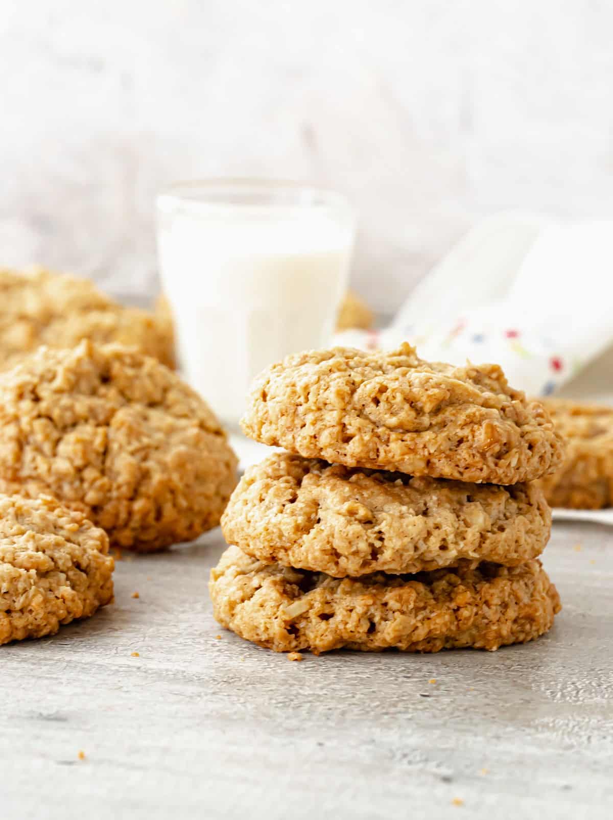 Stack of three oatmeal cookies on a grey surface with glass of milk and other cookies in background.