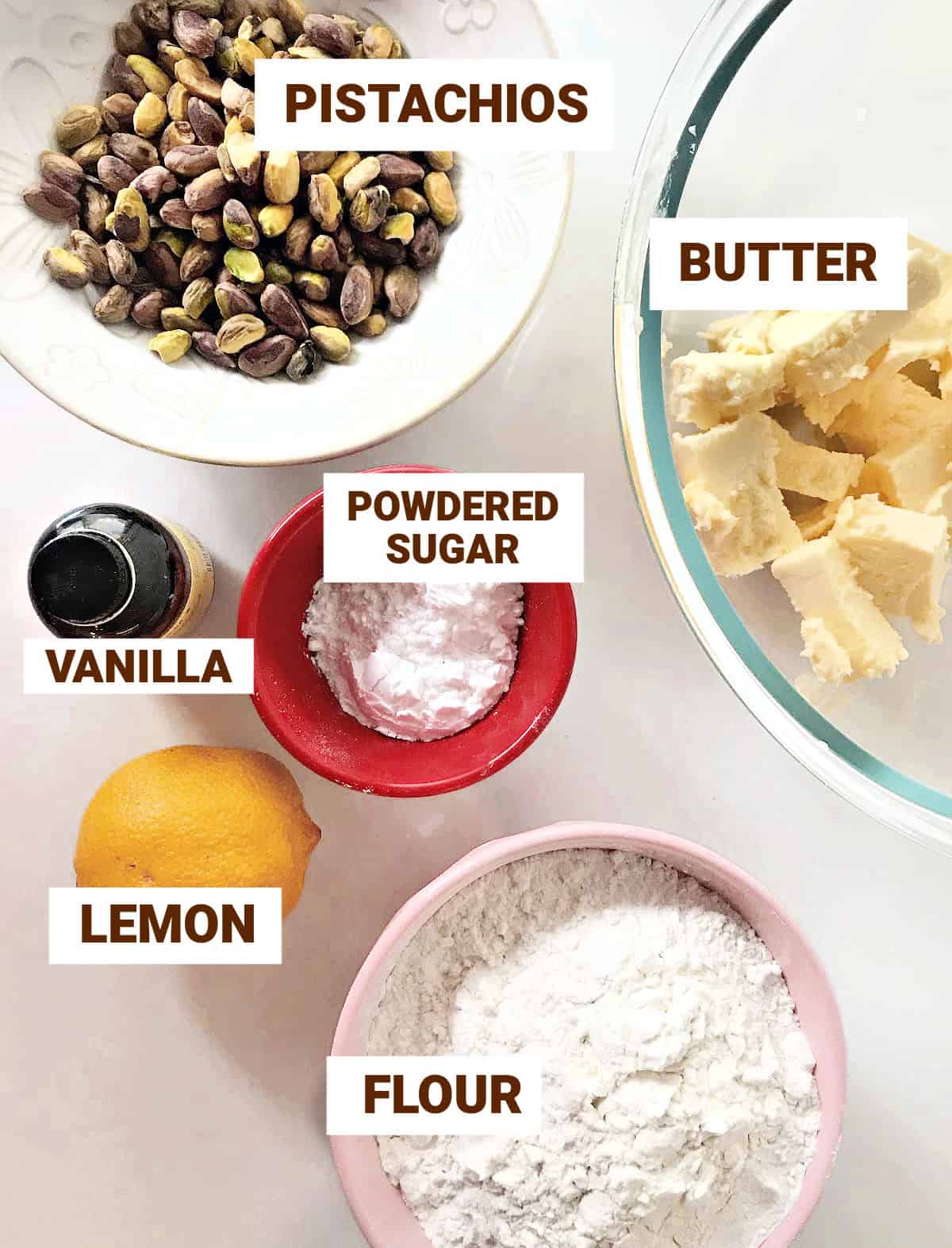 Pistachio lemon cookies ingredients in bowls on a white surface including flour, butter, sugar, vanilla.