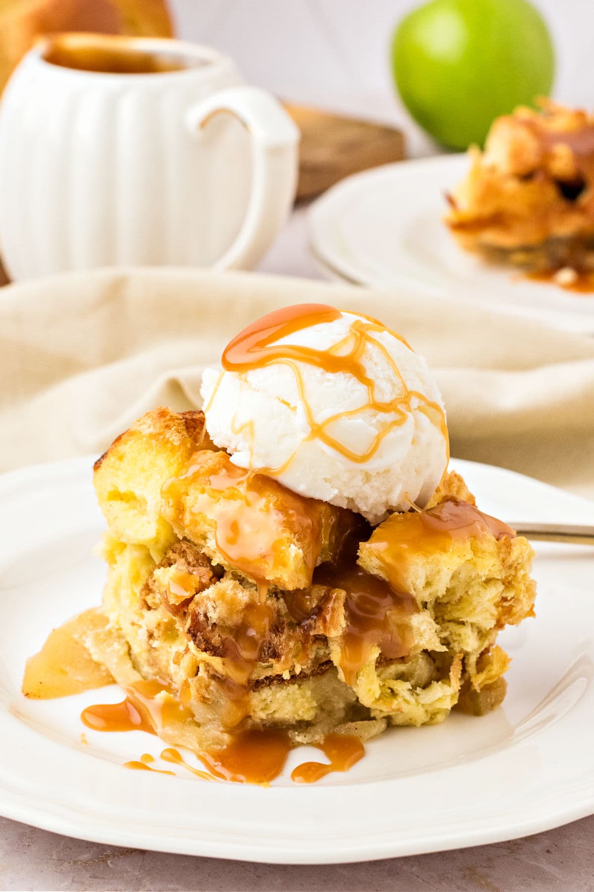 Caramel and cream topped serving of apple brioche pudding on a white plate. White jug, green apple in the gray background.
