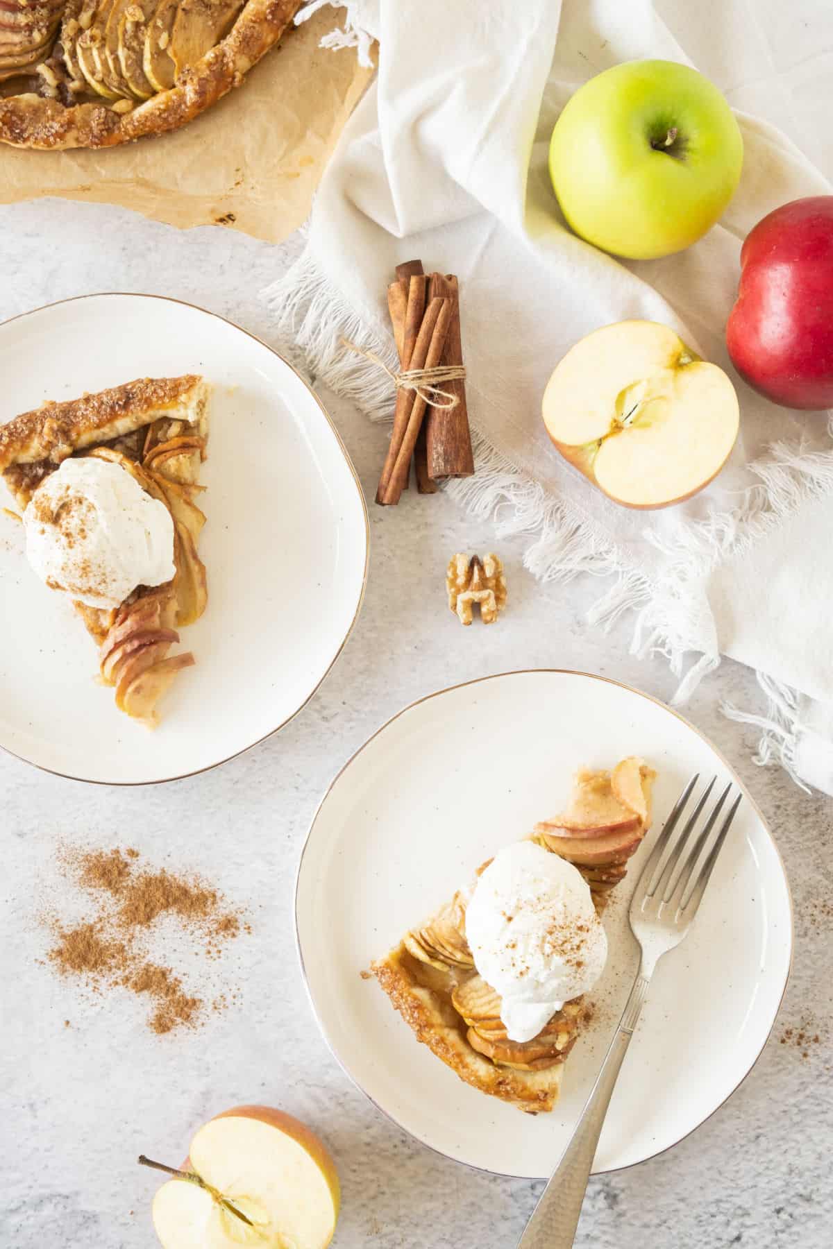 Two white plates with apple galette and cream. Grey surface, fresh apples, cinnamon sticks, walnuts.