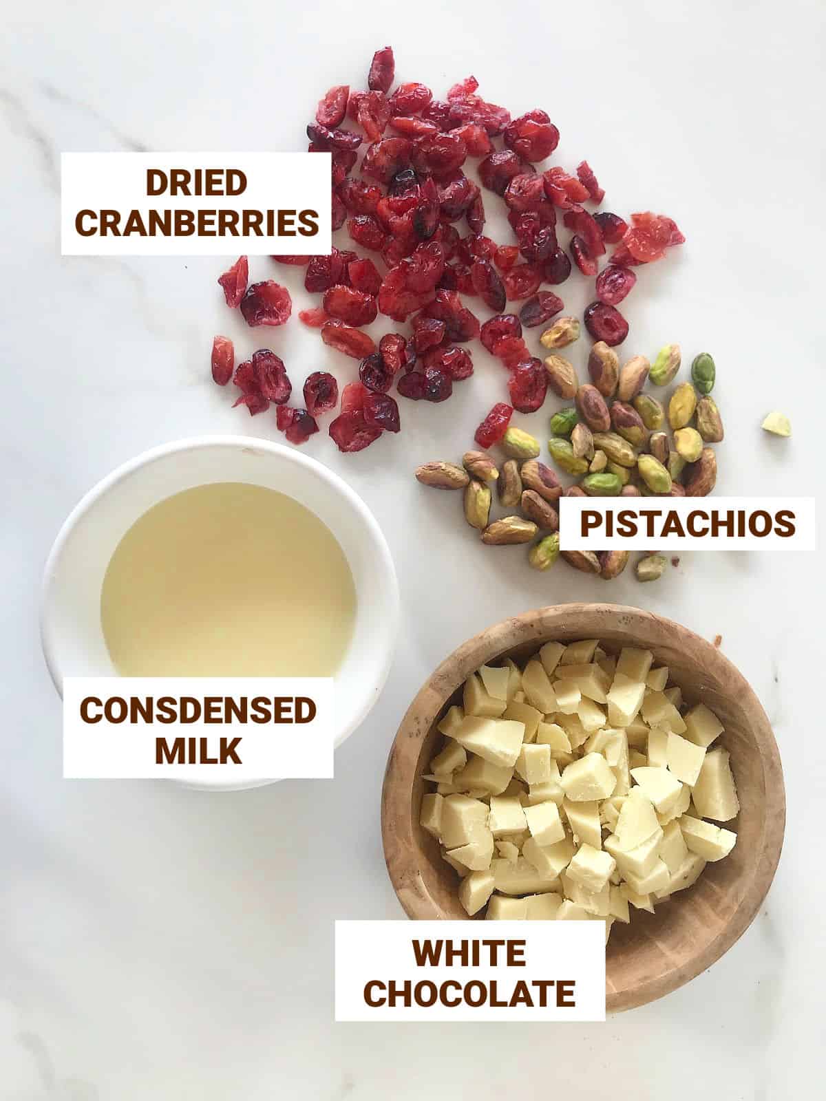 Loose cranberries and pistachios, white chocolate and condensed milk in bowls on a white surface.