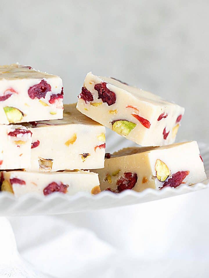 Several pieces of pistachio cranberry white chocolate fudge on a white cake stand with grey background.