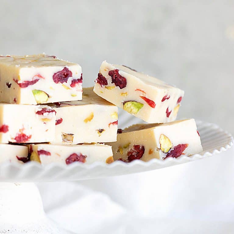 Several pieces of pistachio cranberry white chocolate fudge on a white cake stand with grey background.