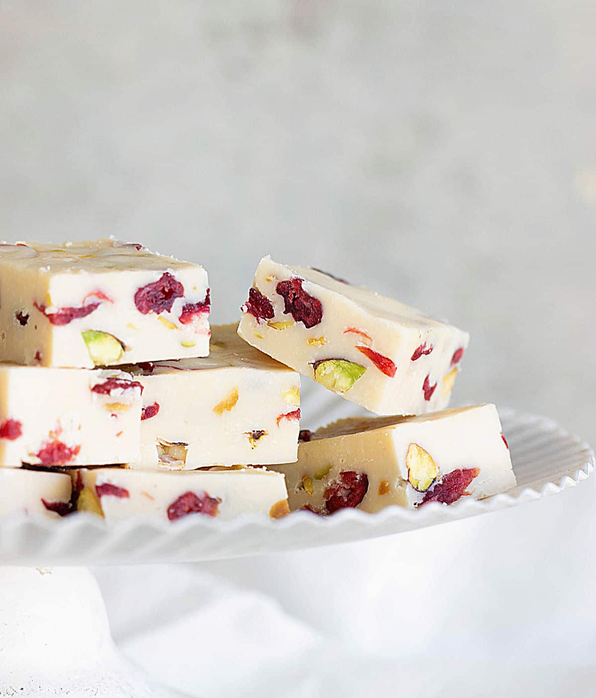 Squares of white fudge with cranberries and pistachios on a white cake stand, light grey background.