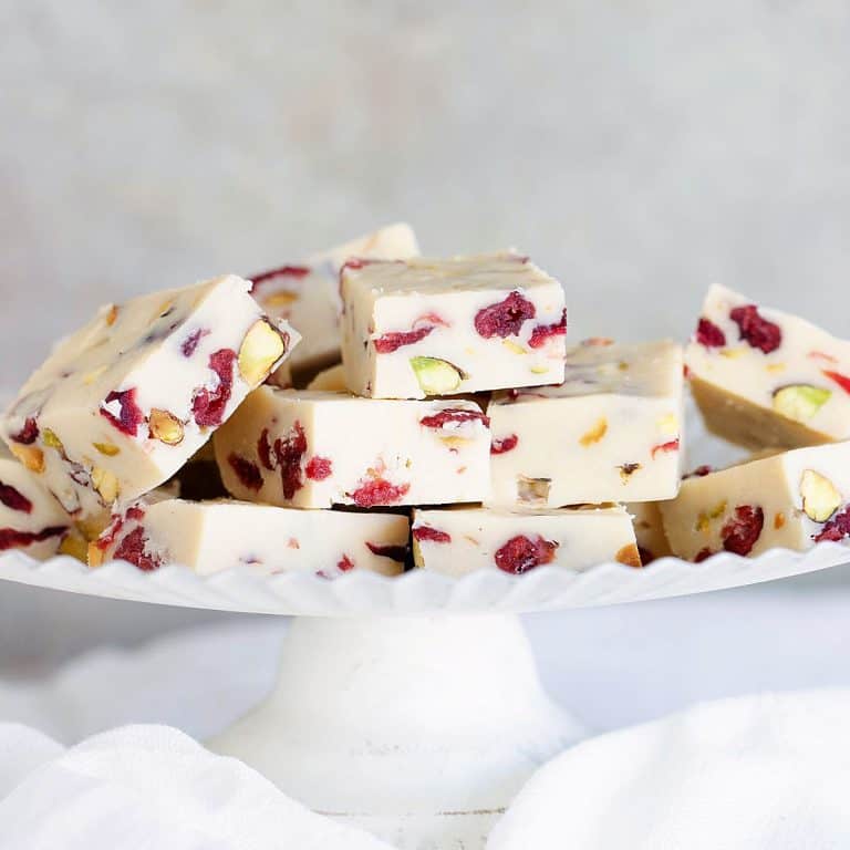 Pile of white chocolate fudge with pistachios and cranberries on a white cake stand. Grey background.