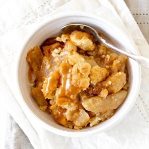 Top view of a white bowl with serving of apple dump cake, a spoon, white marble surface