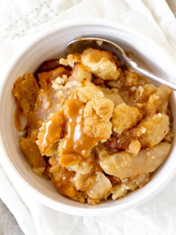 White bowl with serving of fresh apple dump cake with caramel; a silver spoon.