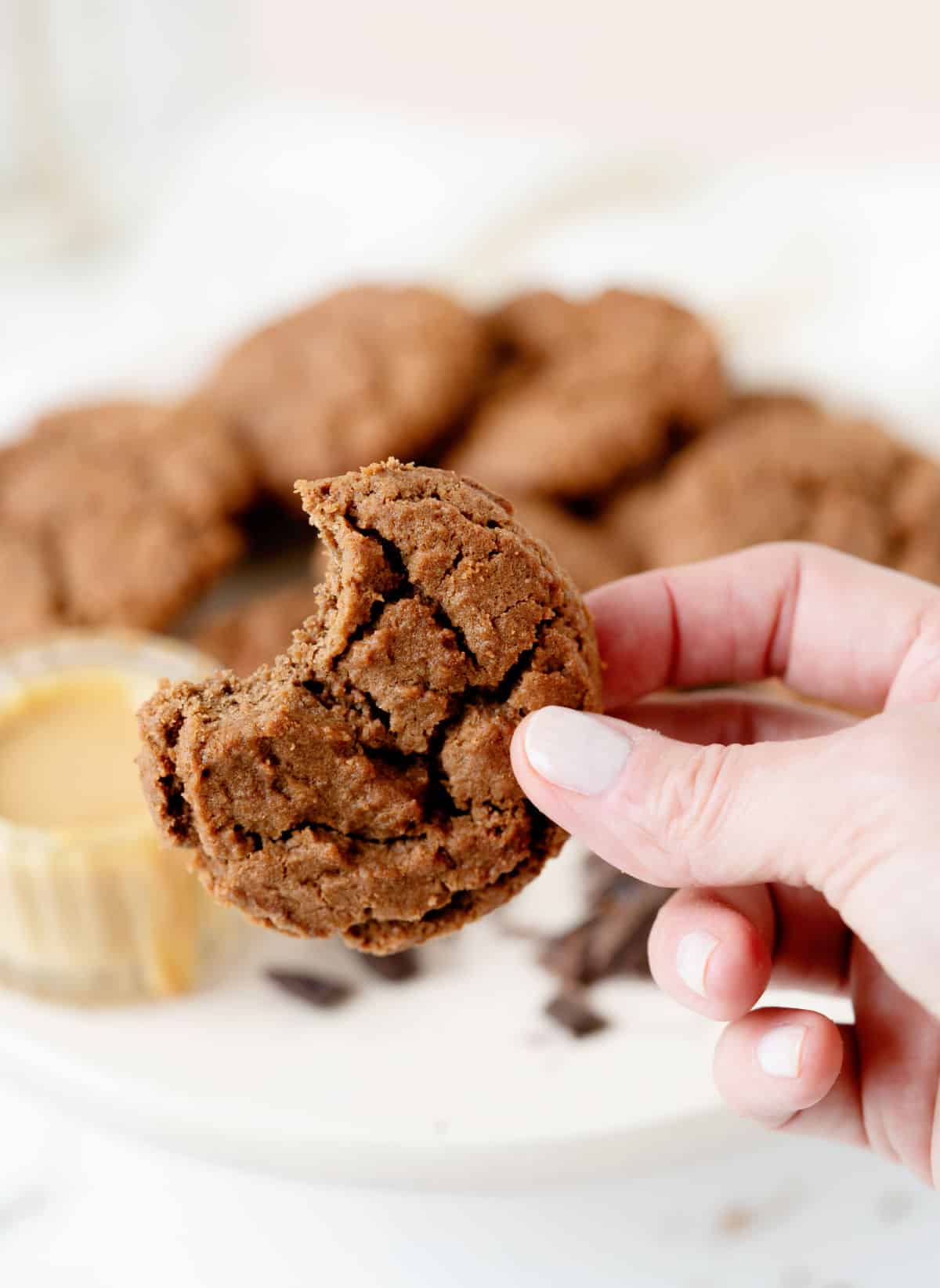 Holding a bitten chocolate cookie with white background and blurred plate of cookies and bowl of peanut butter.