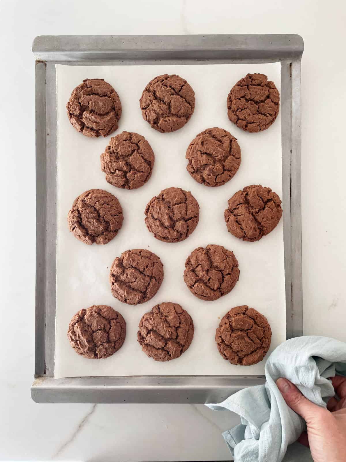 Baked chocolate cookies on a metal pan with parchment paper on a white marble surface. Hand holding it with a towel.
