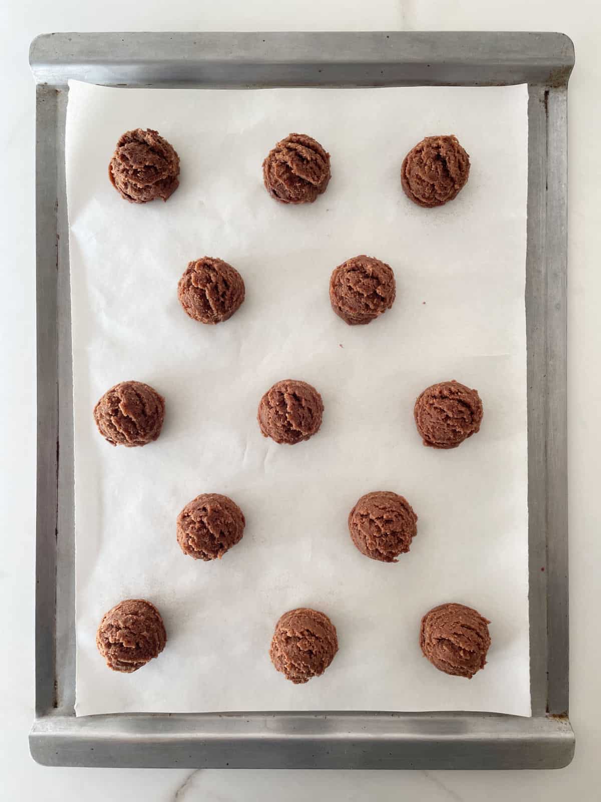Unbaked chocolate cookies on a white parchment paper on a metal sheet on white marble. 