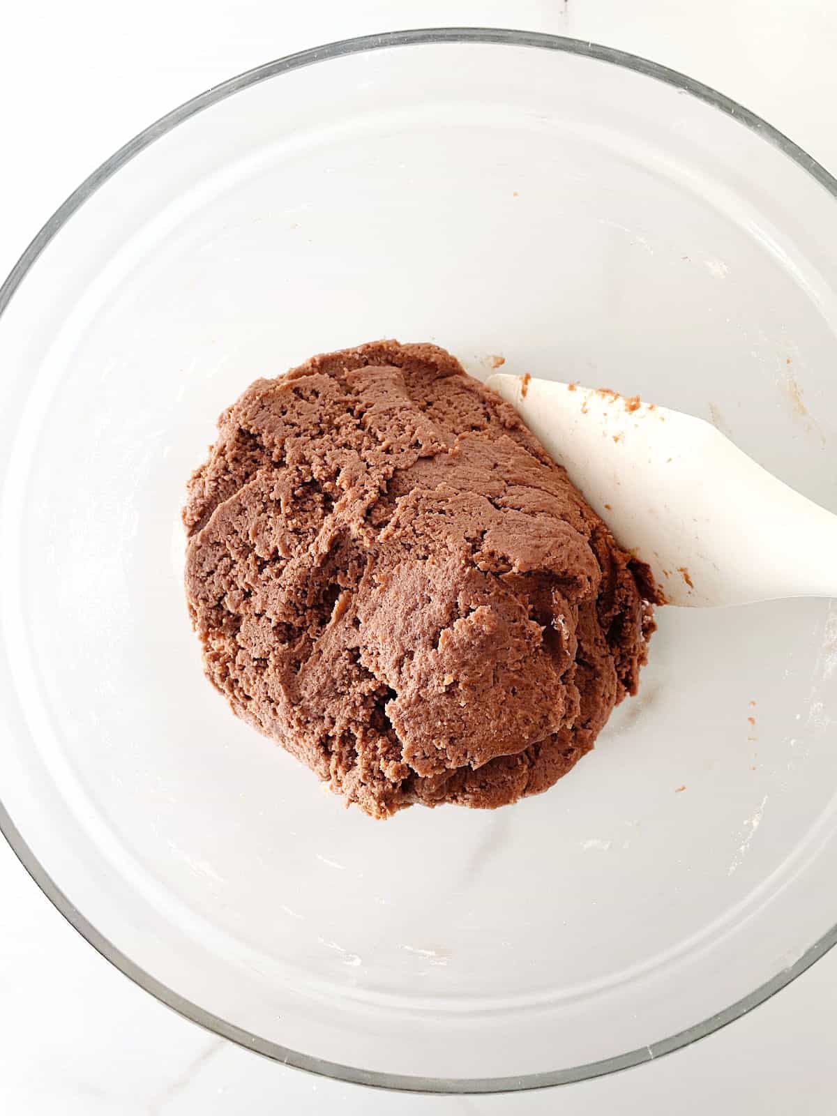 Top view of chocolate cookie dough in a glass bowl with white spatula on a white surface.