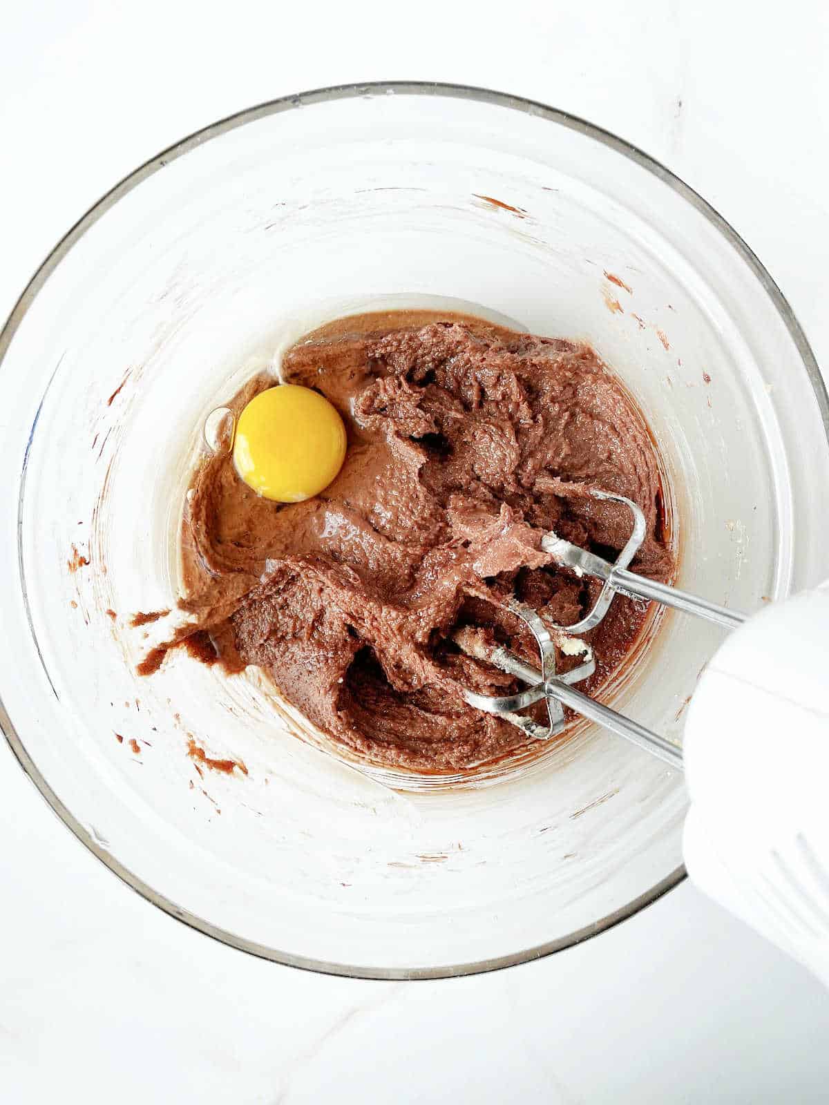 Egg added to chocolate cookie batter in a glass bowl with an electric mixer. White surface.