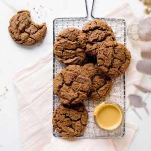 View from above of rectangular cooling rack with pile of chocolate cookies on a white surface, a pink cloth, peanut butter in a bowl.