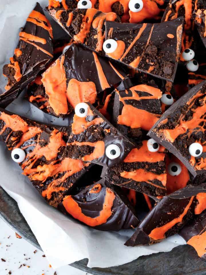 Pieces of dark and orange halloween bark with candy eyes on a metal plate with white paper.