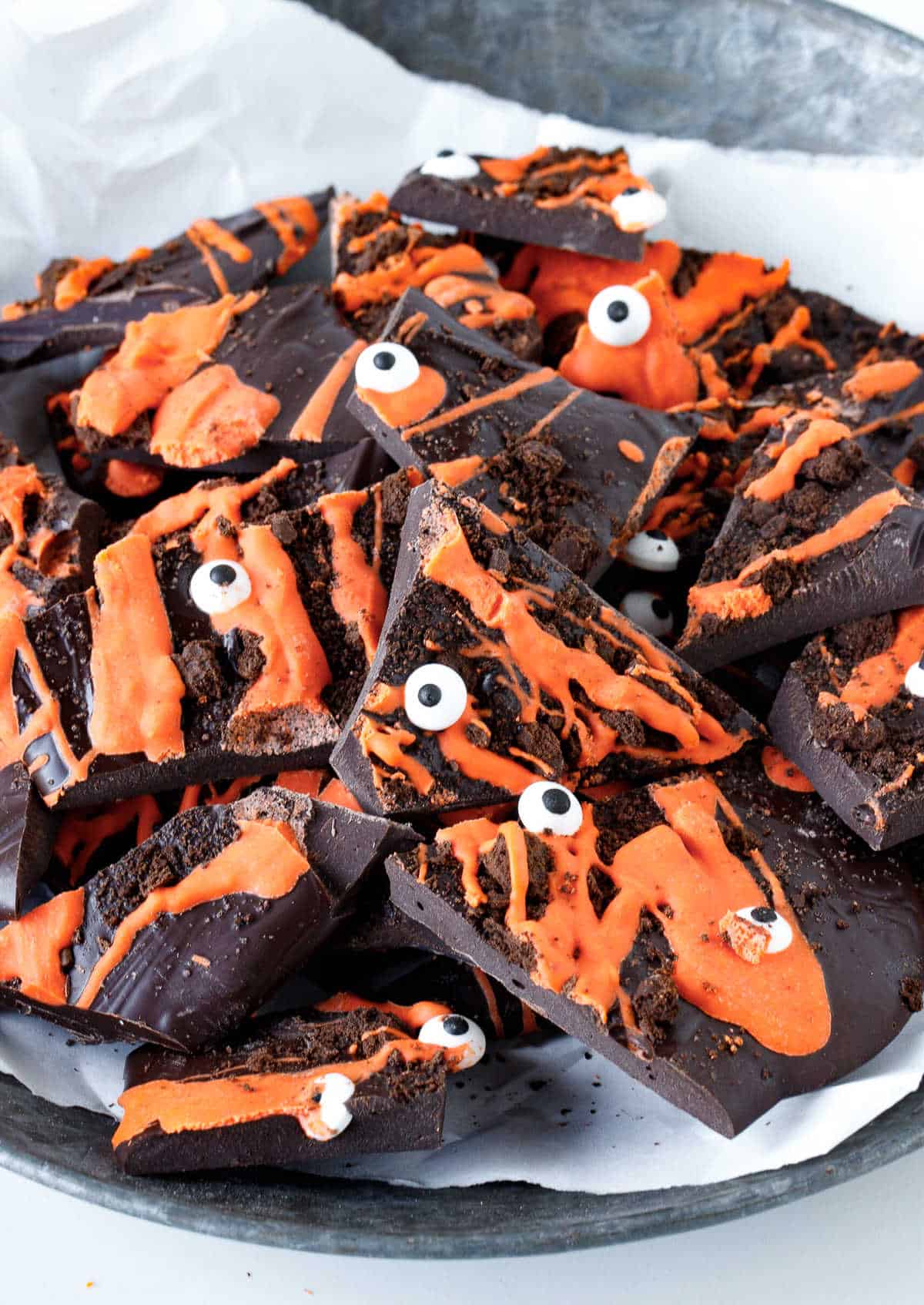 Close up of dark and orange colored chocolate bark in pieces with crushed cookies and candy eyes on a white paper over a metal plate.