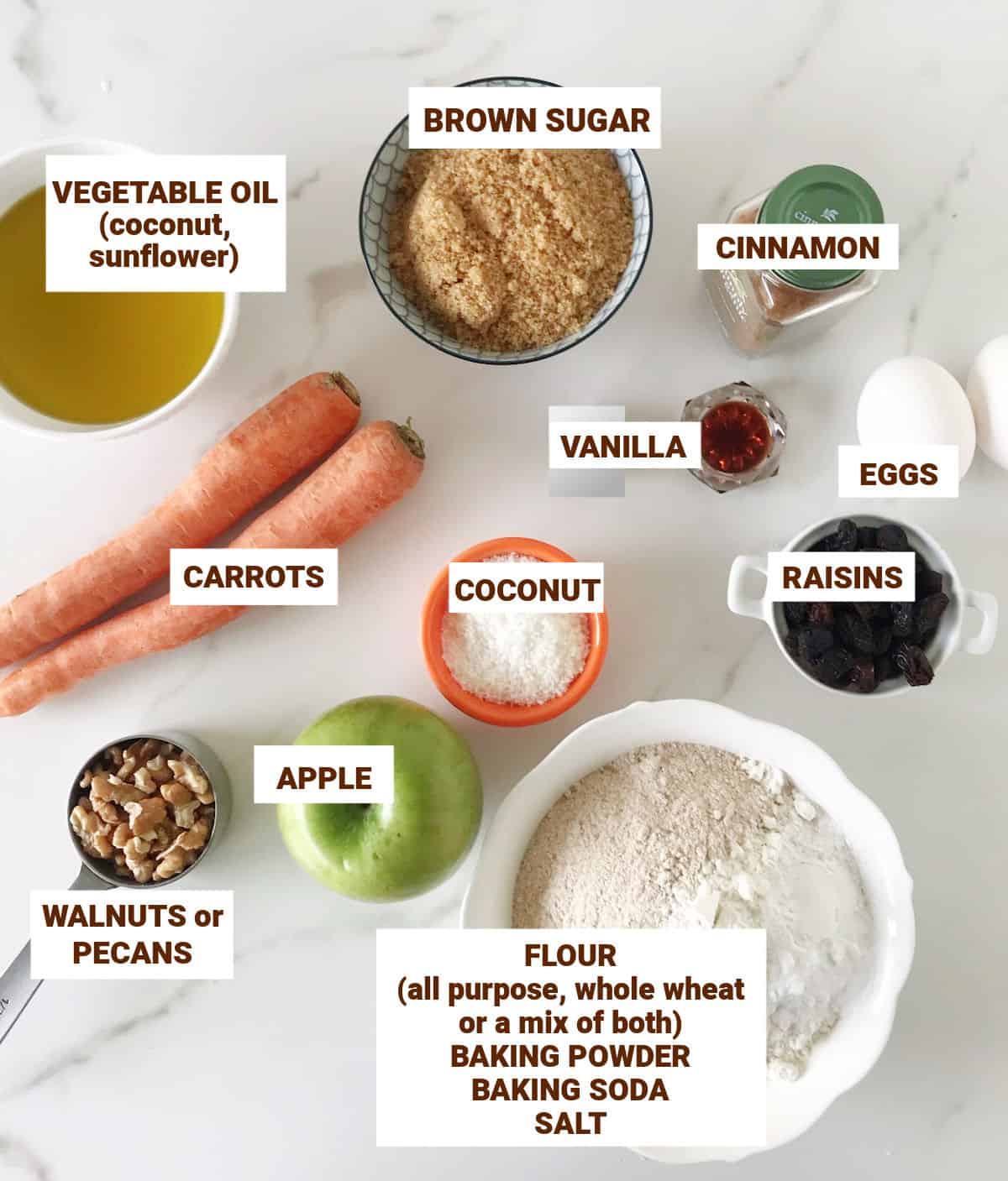 Ingredients in bowls on white marble surface including apple, carrots, oil, spices, walnuts, flour, raisins, sugar.