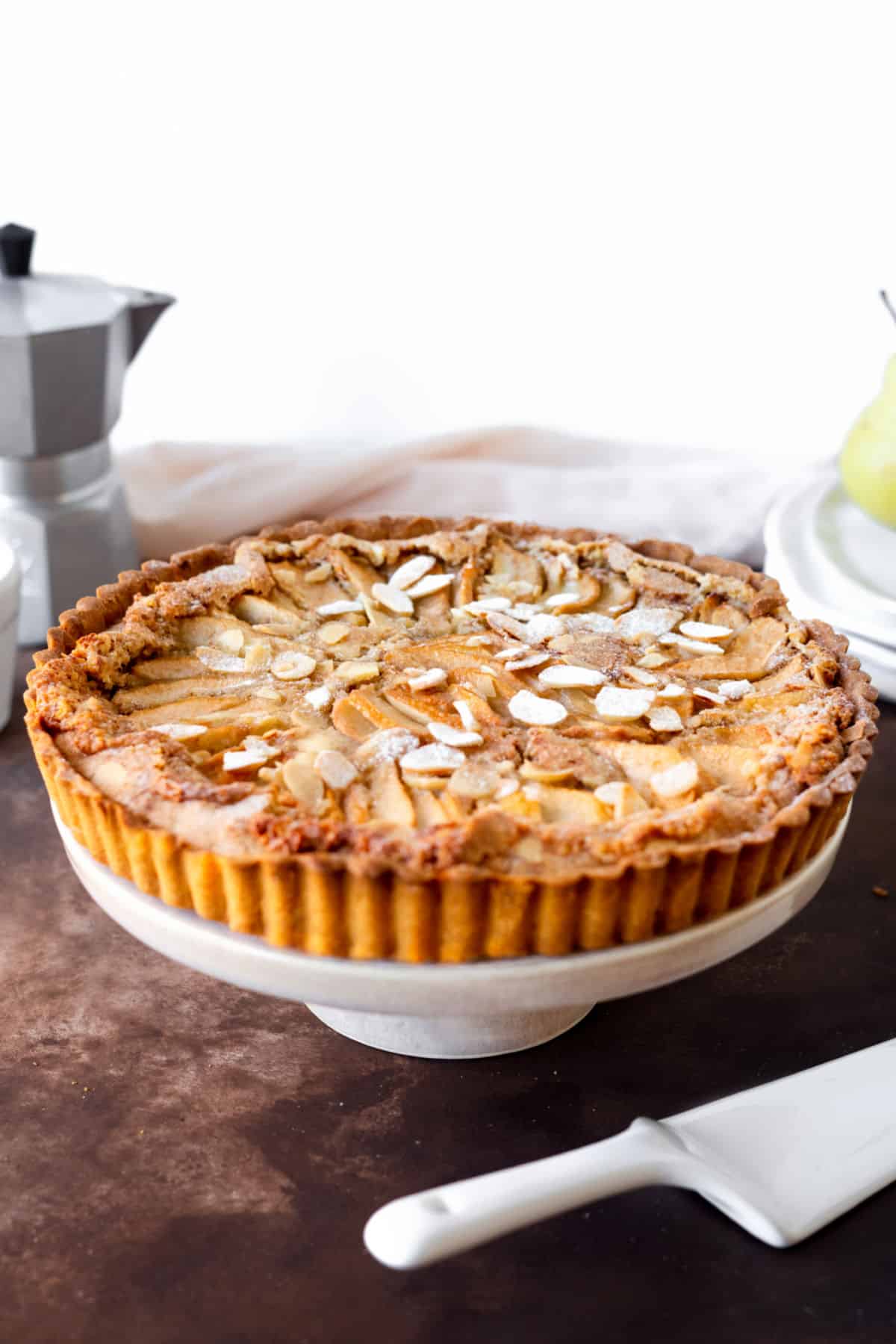 Round pear almond tart on a white cake stand. Brown surface, white background.