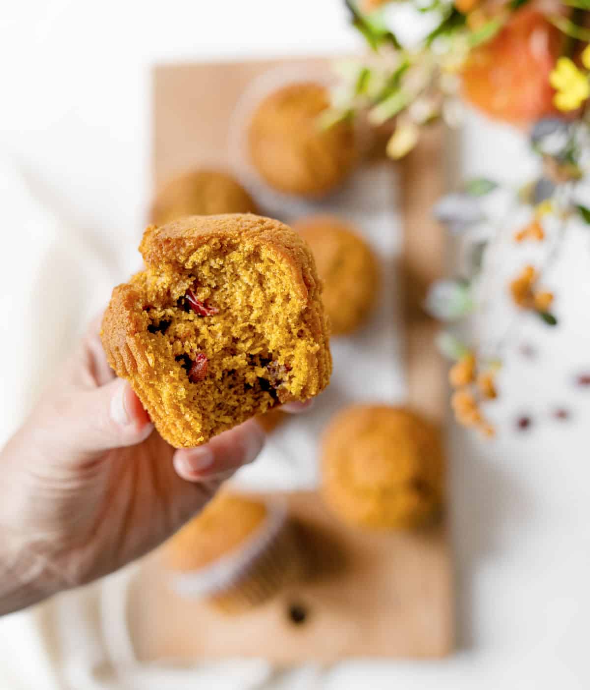 Hand holding eaten pumpkin cranberry muffin over blurred board with more muffins and flower arrangement. 
