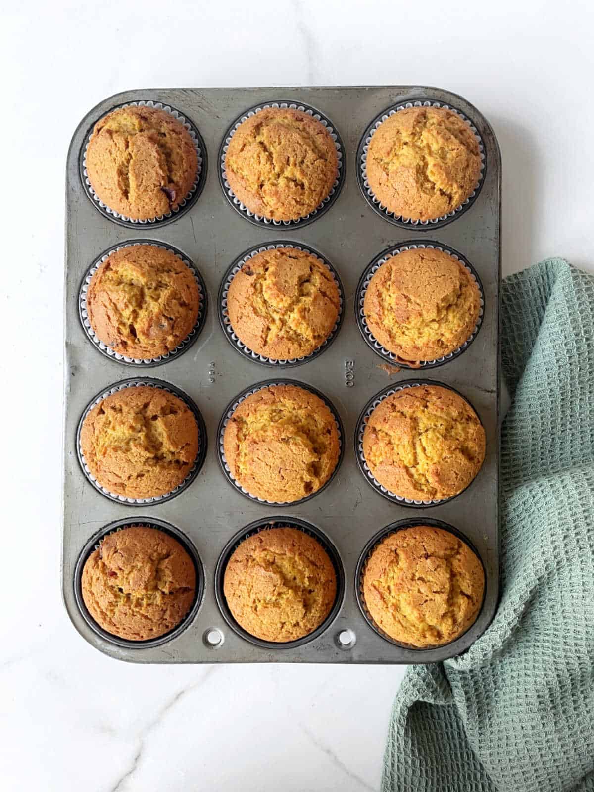Baked pumpkin muffins in the metal pan on a white surface with a green kitchen towel.