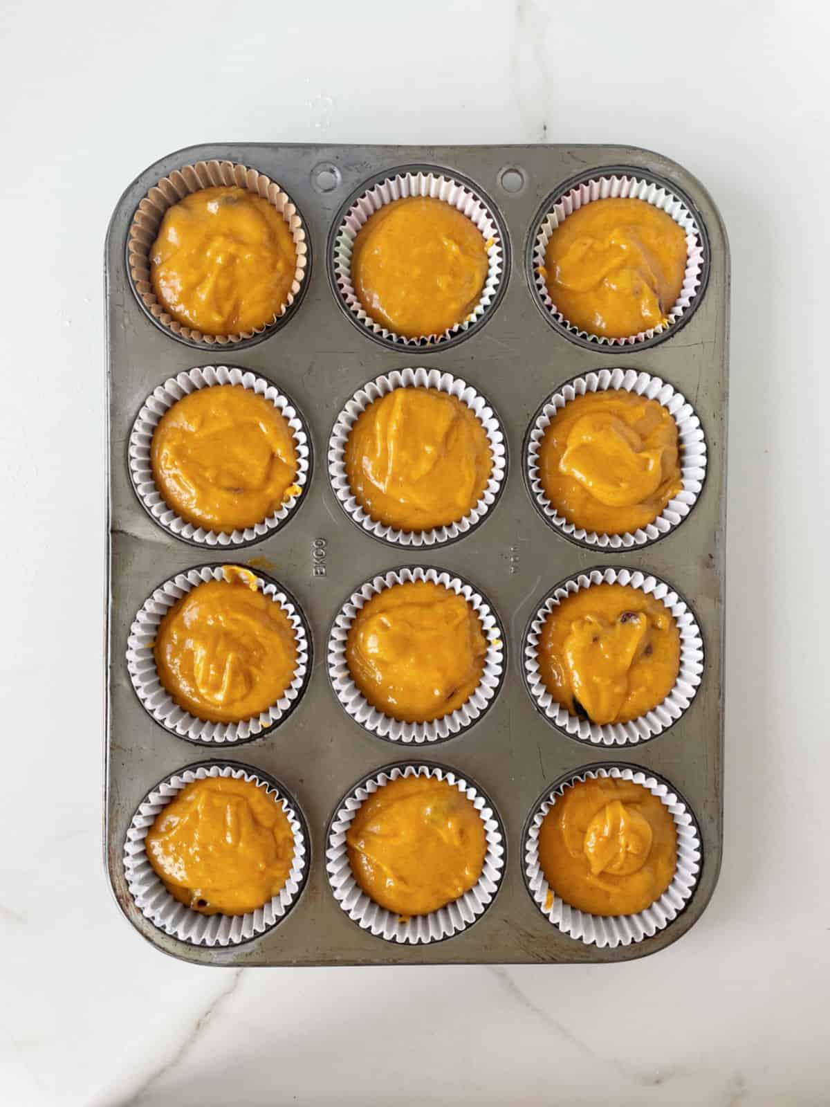 Pumpkin muffin batter in paper liners in a metal pan on a white marble surface.