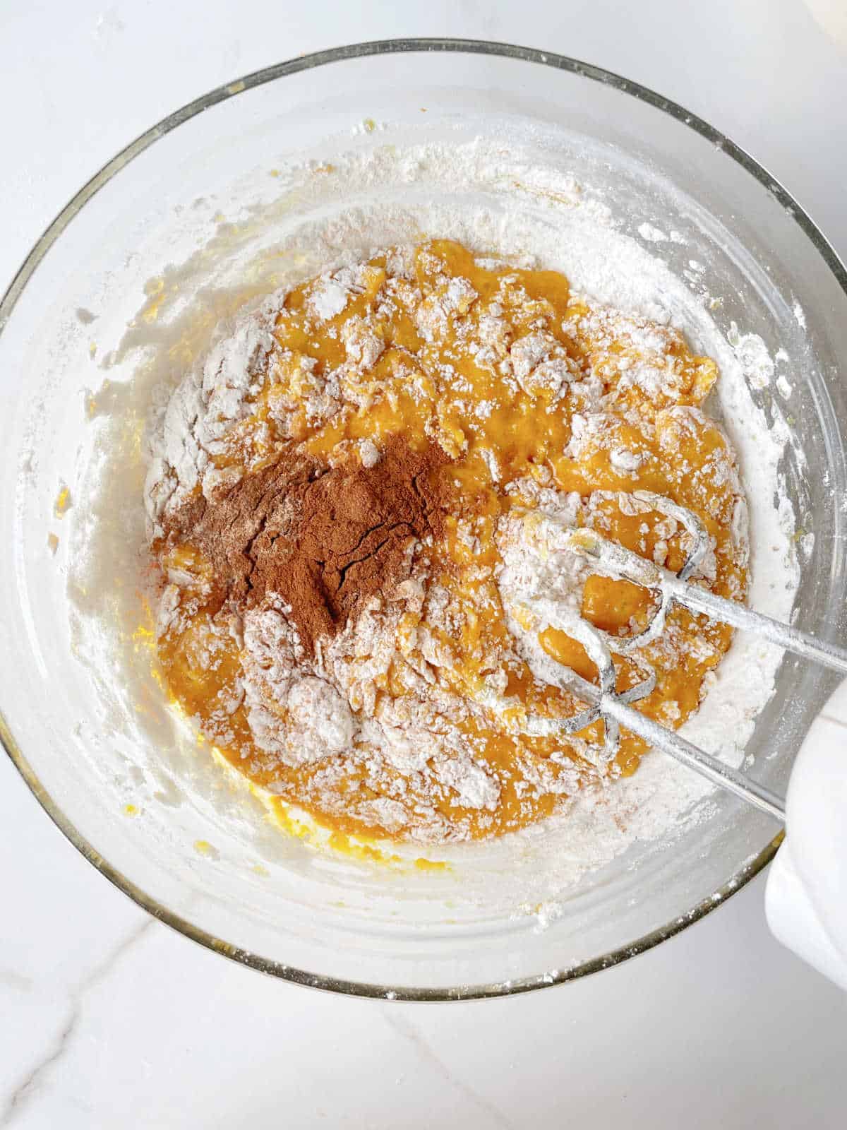 Pumpkin batter with flour and spices in a glass bowl and an electric mixer. White marble surface.