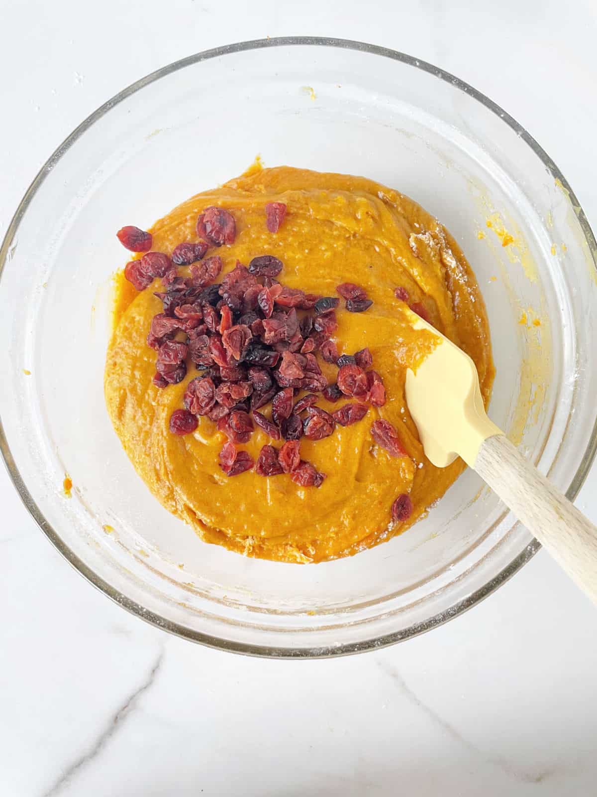 Cranberries added to pumpkin muffin batter in a glass bowl with a yellow spatula on a white marble surface.