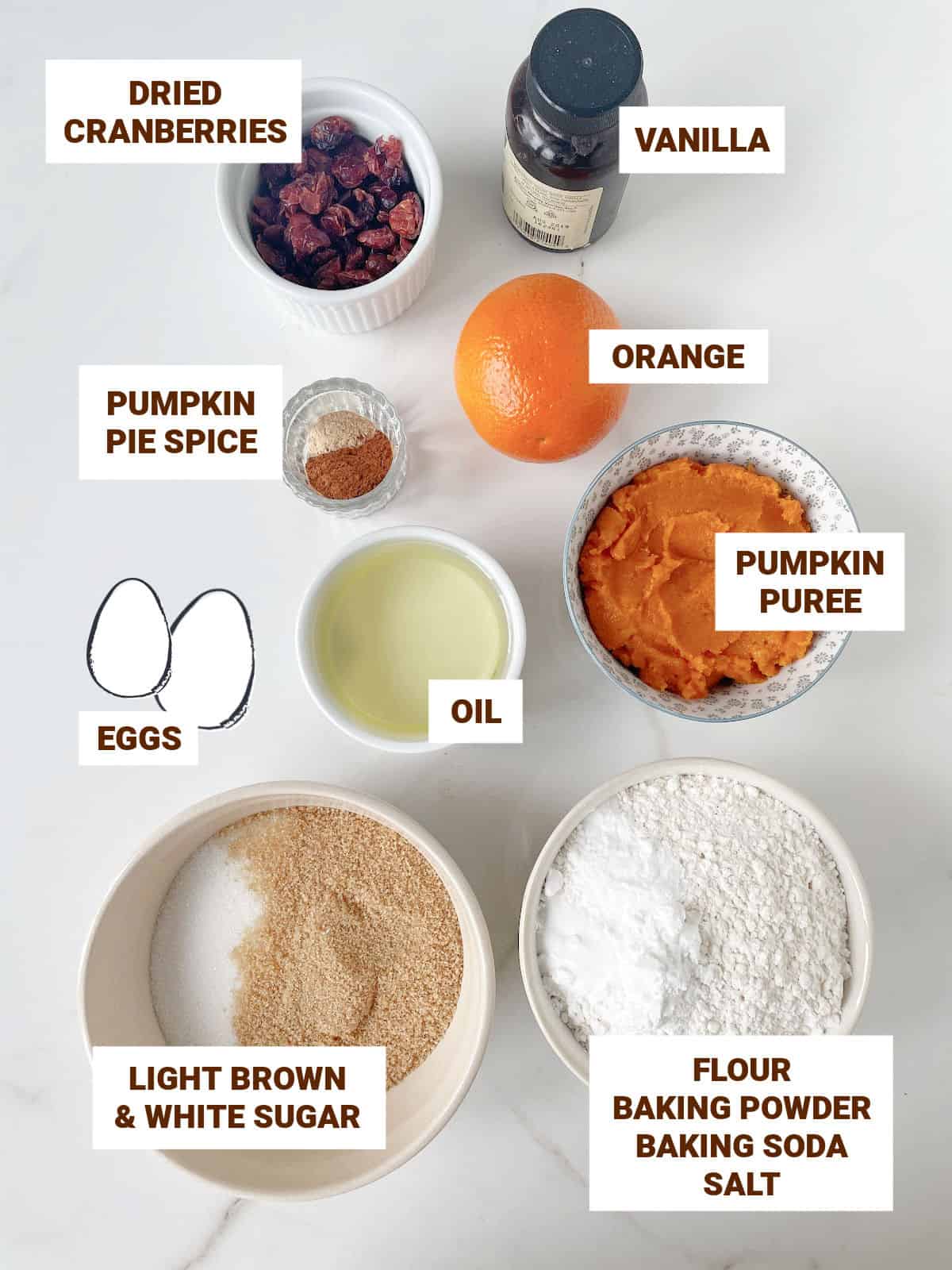 Bowls on white marble surface with ingredients for pumpkin cranberry muffins including oil, eggs, spices, sugar, flour, orange, vanilla.
