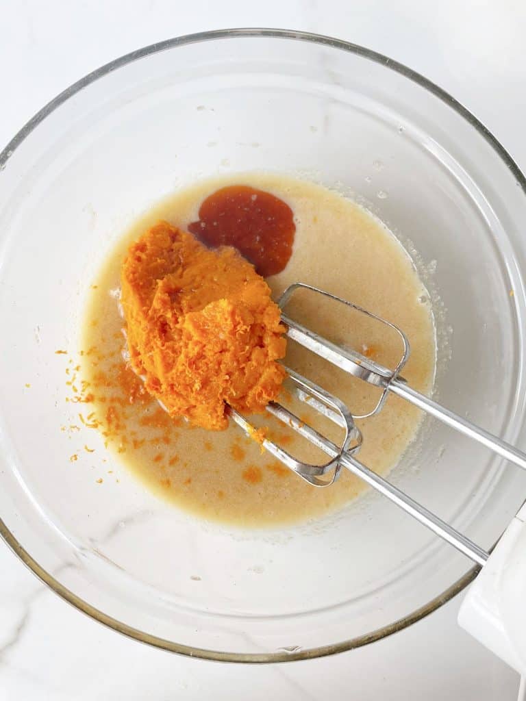 Pumpkin puree, vanilla and oil mixture in a glass bowl with electric mixer. White surface.
