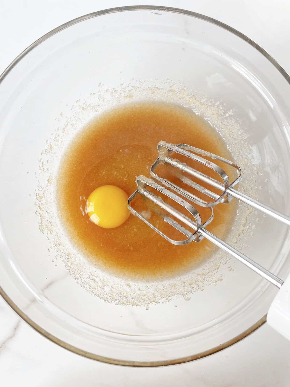 Oil, sugar, and egg on a glass bowl with electric mixer on a white marble surface.