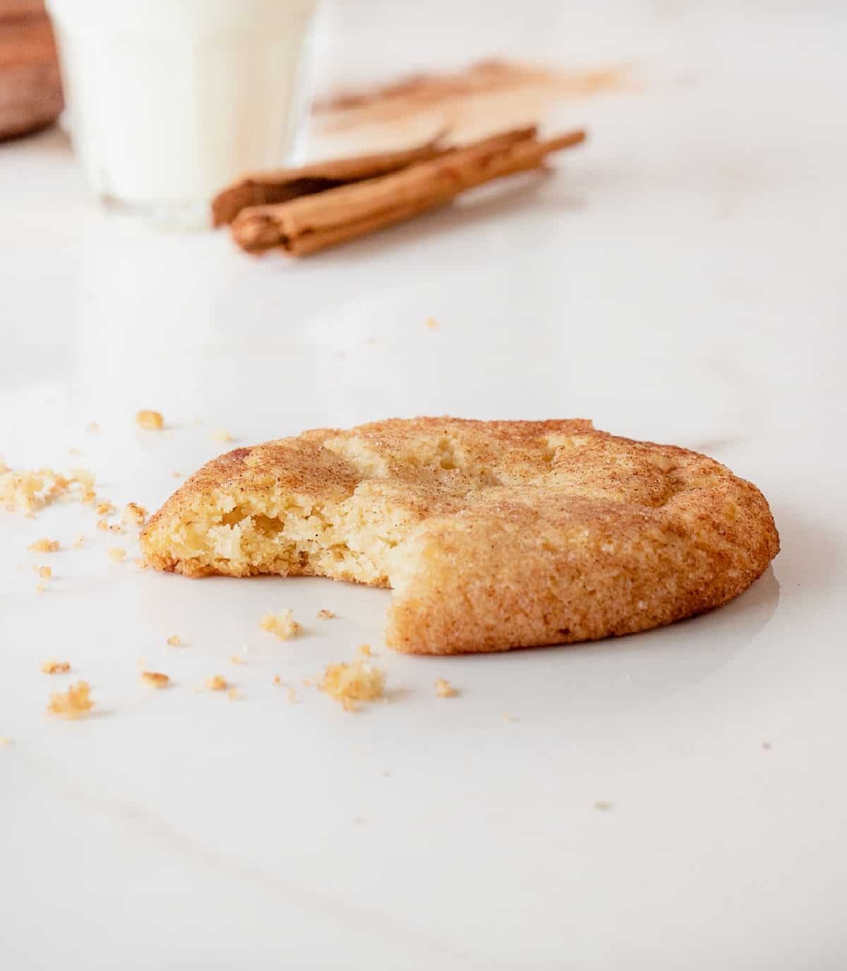 Single bitten snickerdoodle cookie on white marble surface. Cinnamon stick and glass of milk in the background.