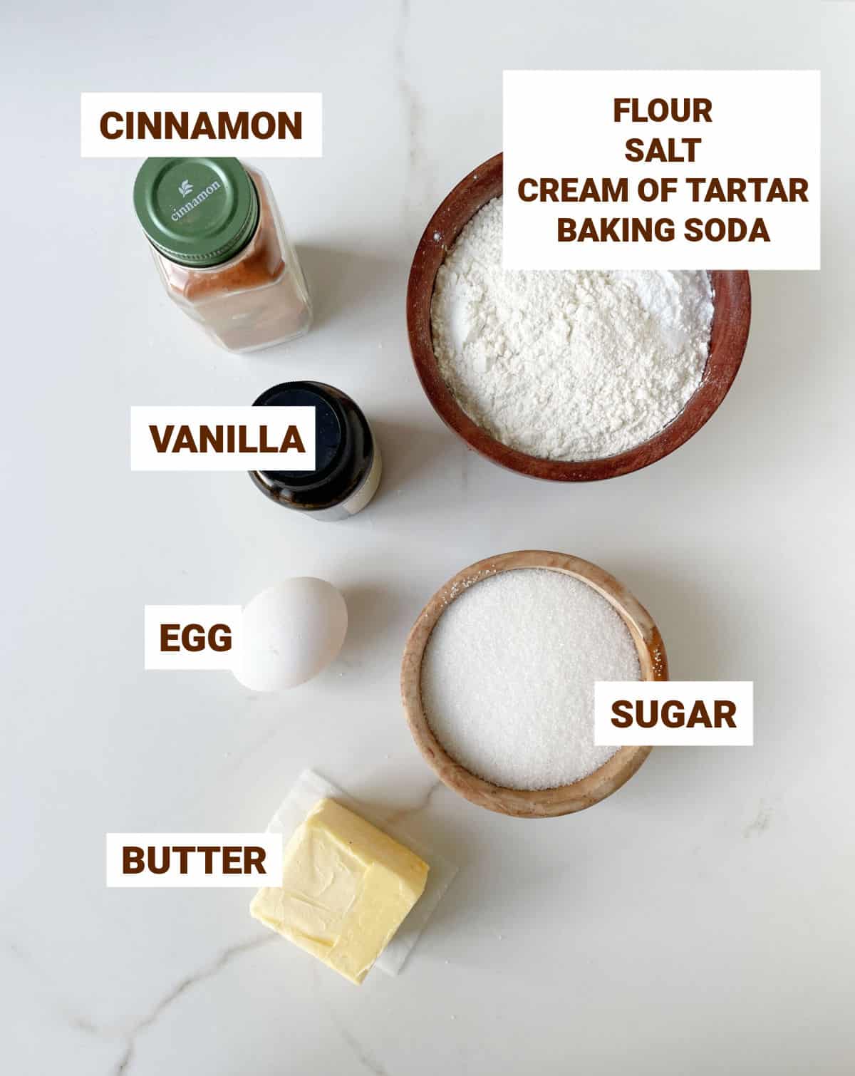 White marble surface with ingredients for snickerdoodles including cinnamon, butter, flour mixture, vanilla, sugar, and egg.