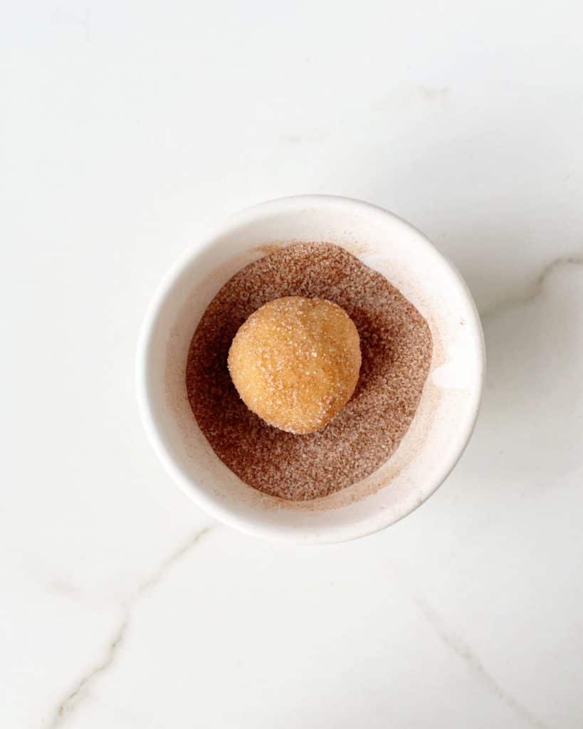 Snickerdoodle cookie ball coated in cinnamon sugar in a white bowl on a white marble surface.