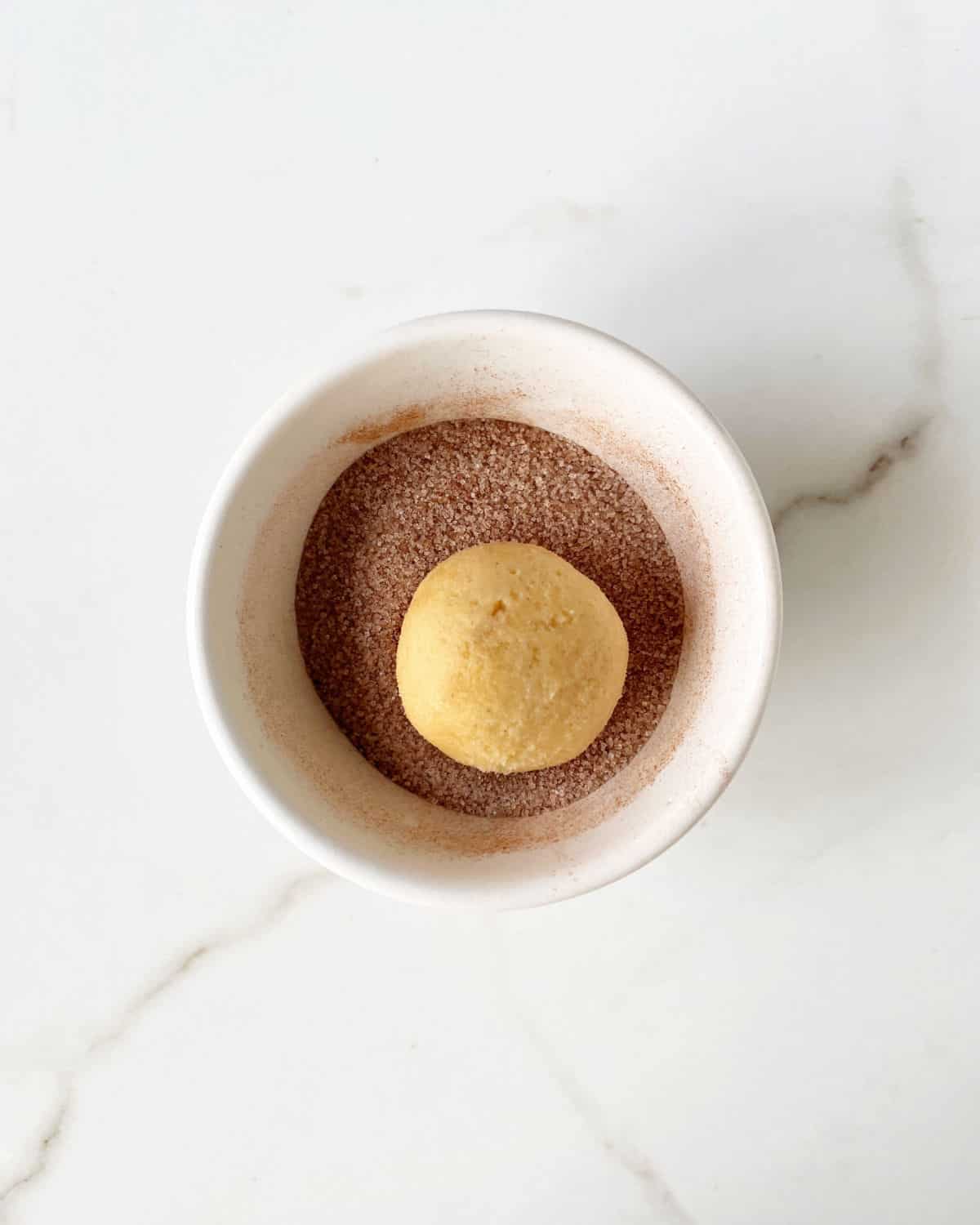 Ball of snickerdoodle cookie dough on a white bowl with cinnamon sugar. White marble surface.