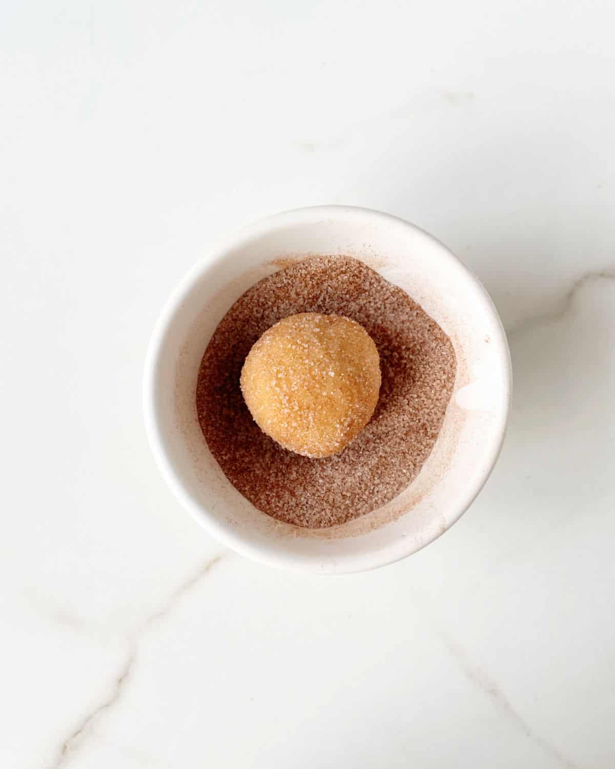Snickerdoodle cookie ball coated in cinnamon sugar in a white bowl on a white marble surface. 