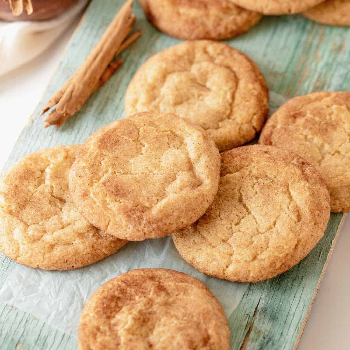 Green wooden board with several snickerdoodles piled and a cinnamon stick.