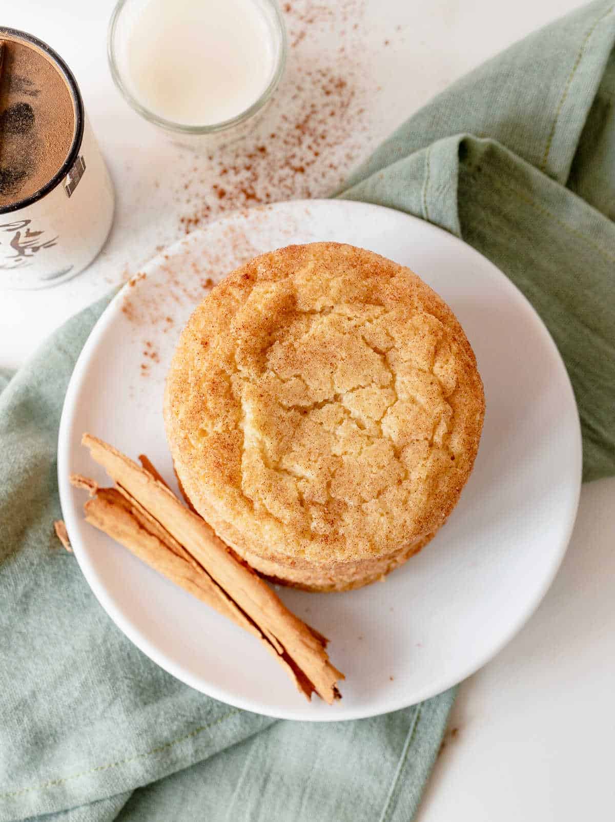 Top view of snickerdoodle cookies on a white plate with a cinnamon stick. Green cloth underneath, white surface.