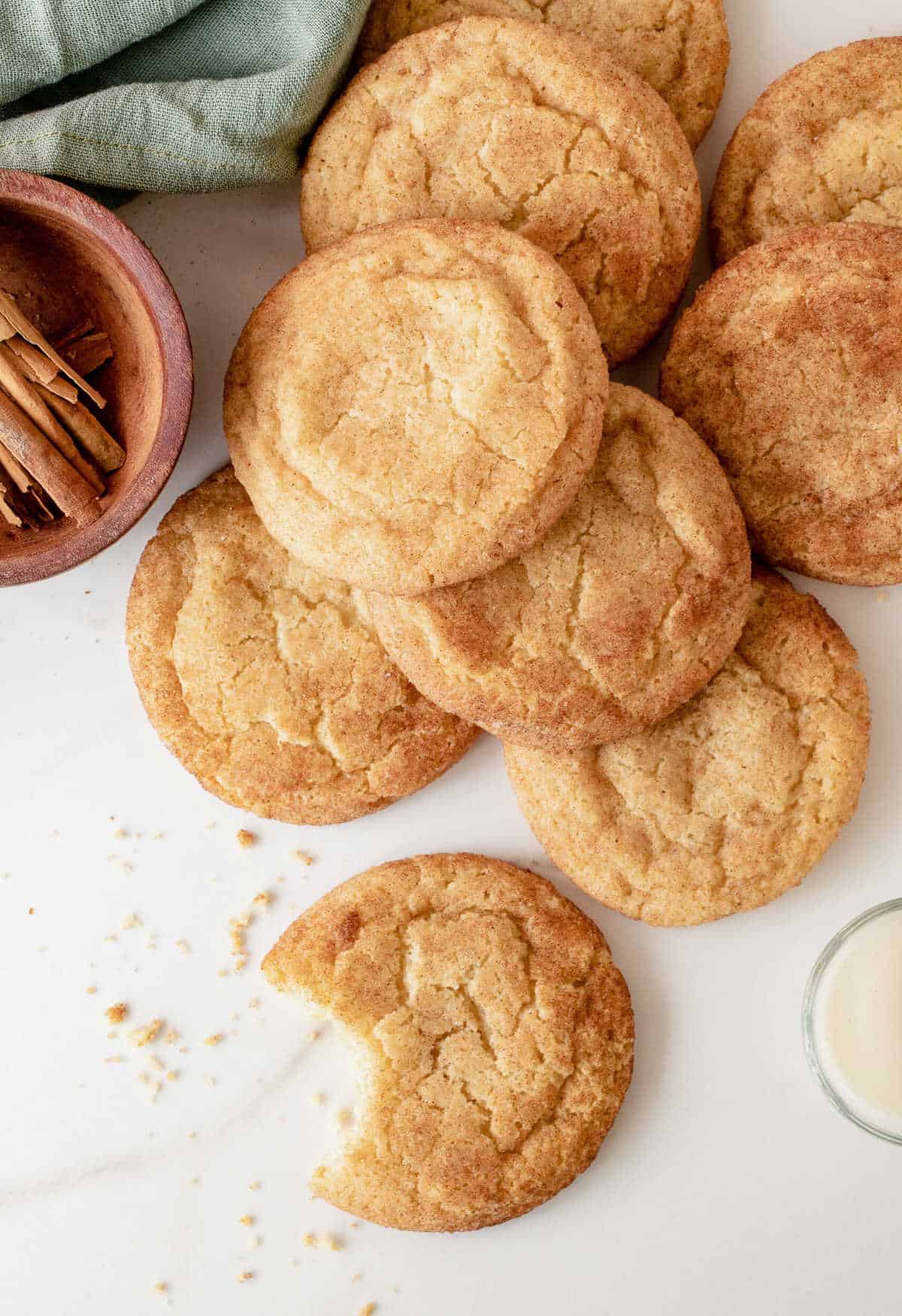 A pile of snickerdoodles and a bitten one on white marble surface with a green cloth, and bowl with cinnamon sticks. View from above.