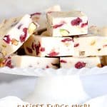 Piled up pieces of white fudge with cranberries and pistachios on a white cake stand. Grey background and text overlay.