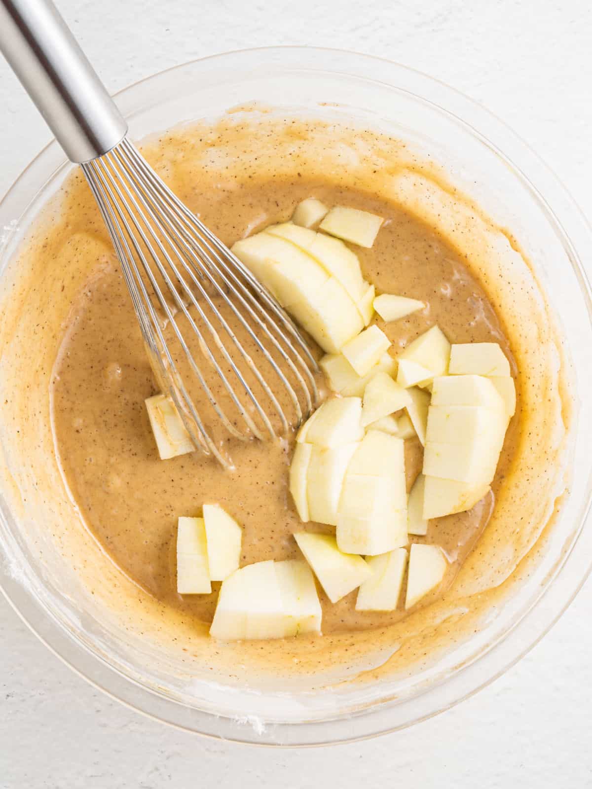 Cake batter with chunks of apple in a glass bowl with a whisk. White surface.