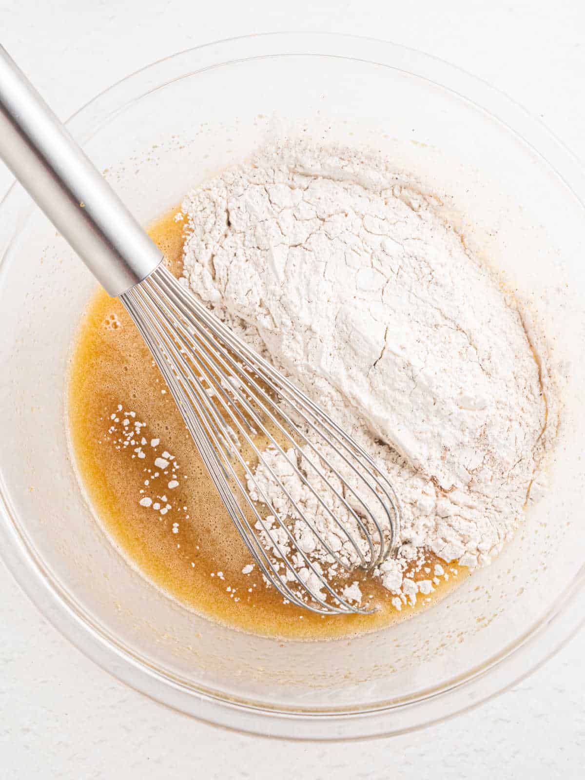 Flour added to cake batter in a white bowl with a whisk inside. White surface.