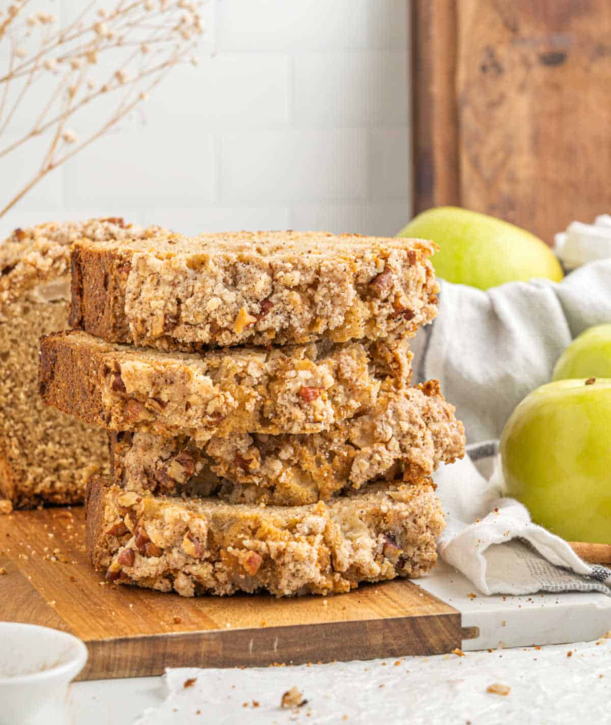 Four slices of apple crumb bread stacked on a wooden board. Green apples and a cloth as props.