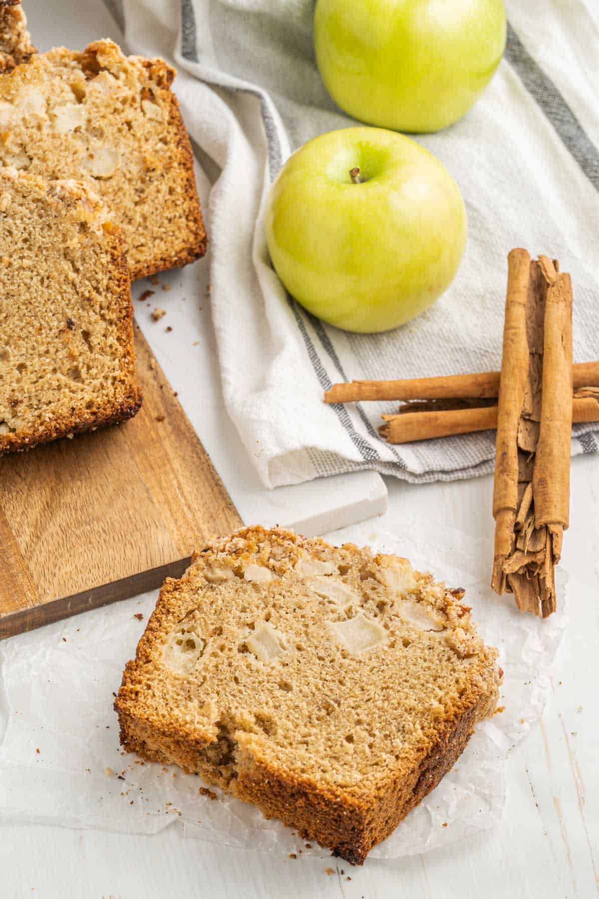 Top view of apple bread slices, a wooden board, green apples, cinnamon sticks, and kitchen cloth.