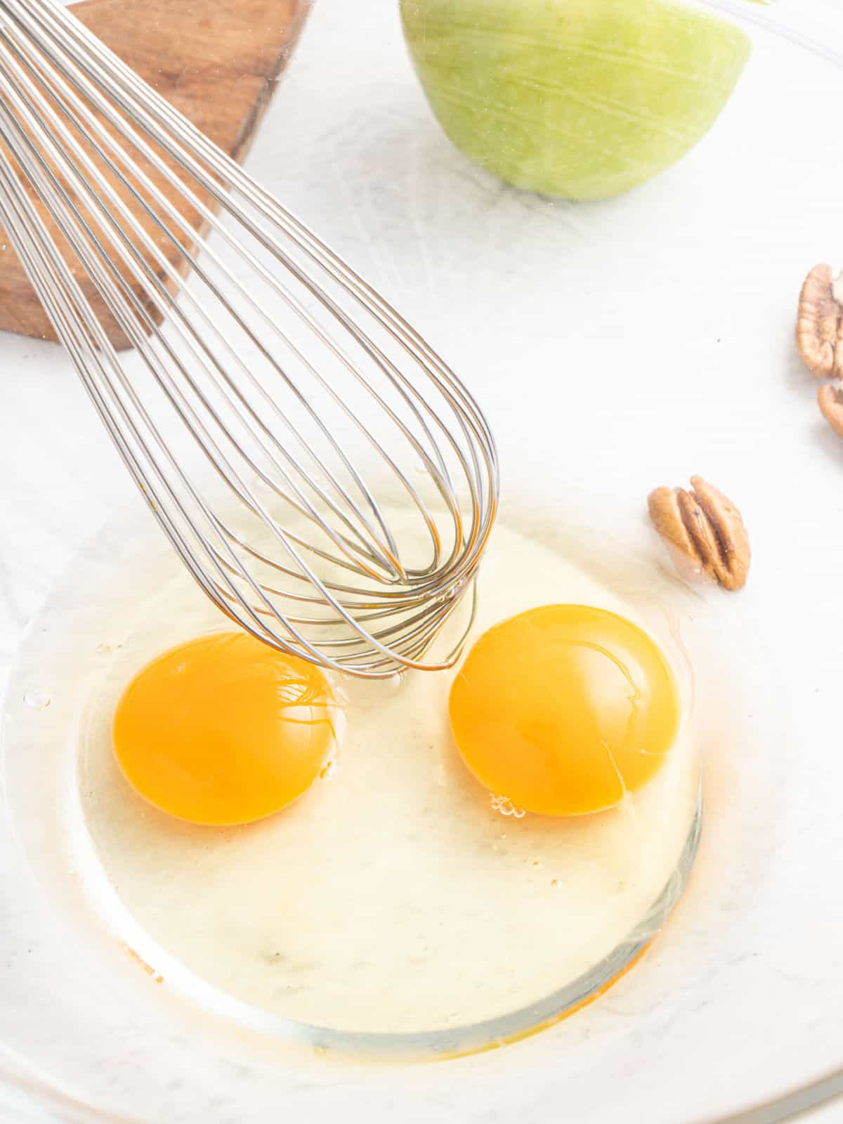 Eggs and oil in a glass bowl with a whisk on a white surface.