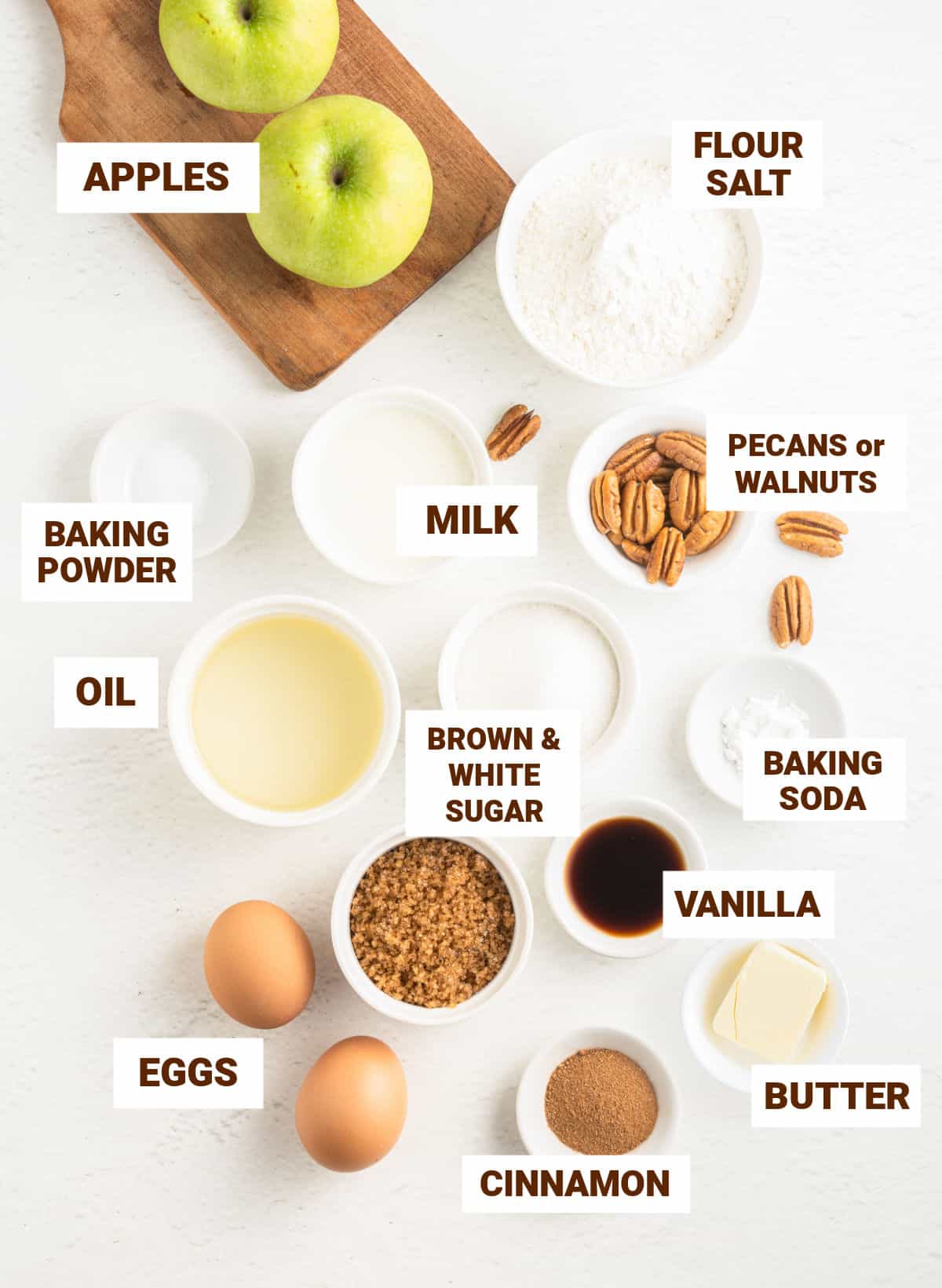 Bowls with ingredients for apple cinnamon bread on a white surface including butter, oil, flour mixture, eggs, sugars, vanilla, pecans, milk.