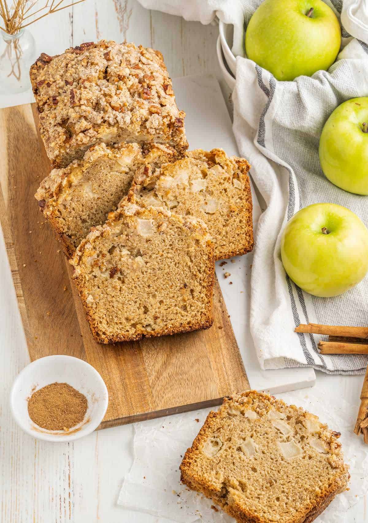 View from above of wooden board on a white surface with apple bread slices, a kitchen cloth, green apples, cinnamon sugar in a bowl.