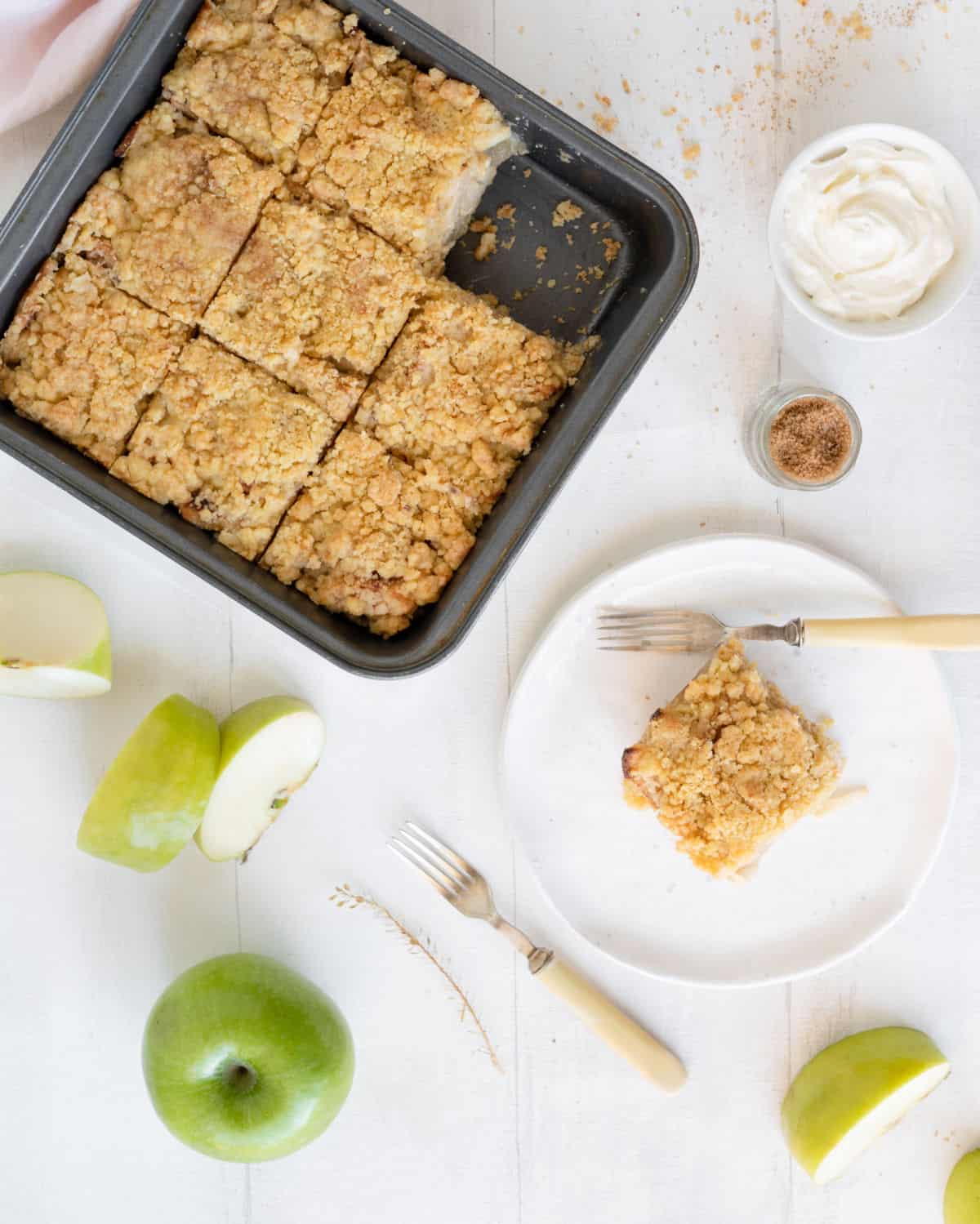 Top view of square metal pan with apple crumb bars, serving on a white plate, white surface, apples and forks around.