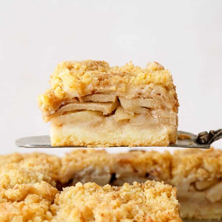 Close up of apple pie crumb bar on a metal cake server. Pink background with more bars.
