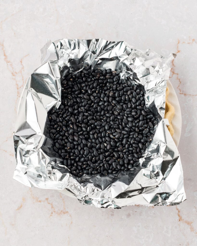Foil and black bean topped unbaked pie crust. White marbled surface.