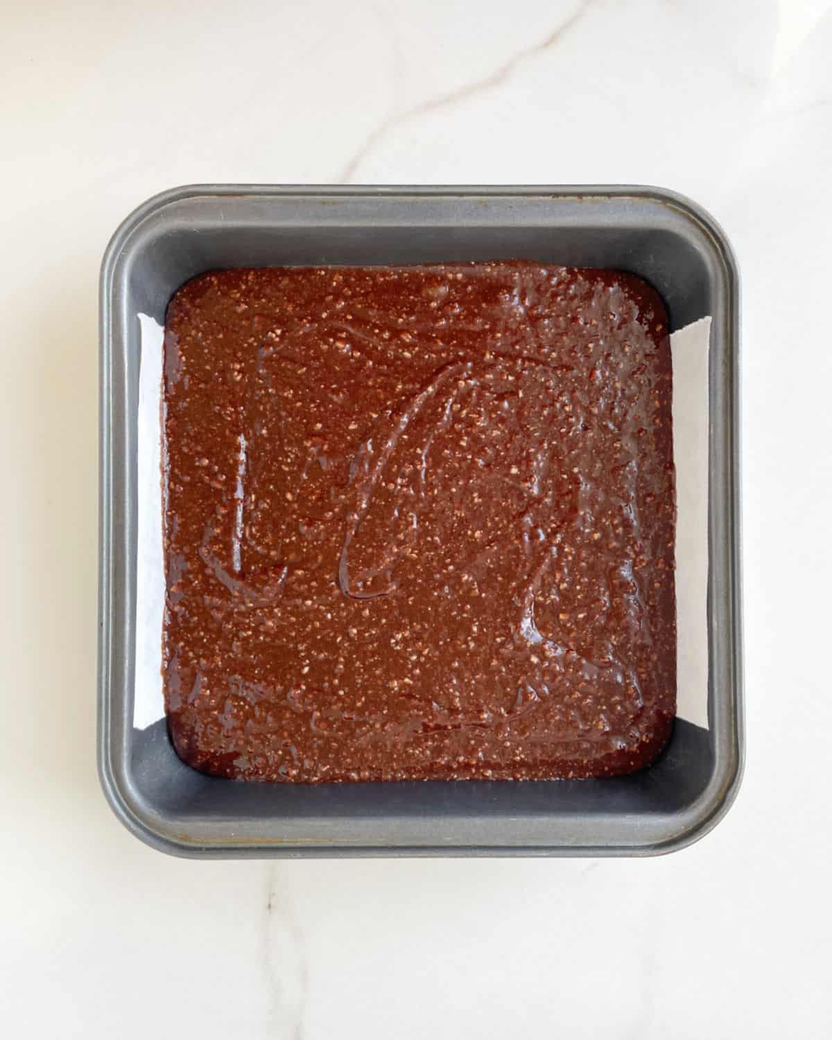 Brownie batter in a square metal pan on a white marble surface.