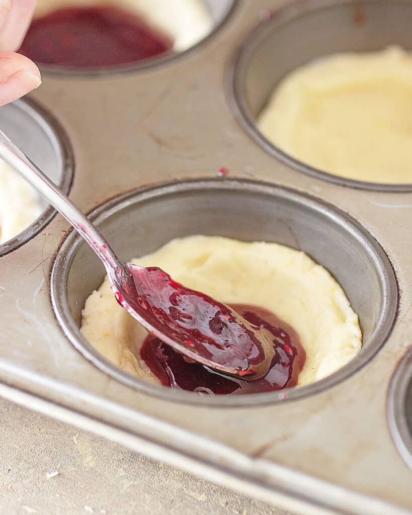 Filling cookie dough cups in a muffin pan with raspberry jam.